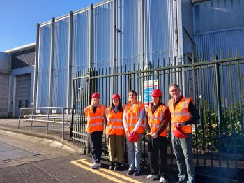 Sussex staff and Community Ambassadors stand in front of the Materials Recovery Facility with high visibility jackets and helmets on