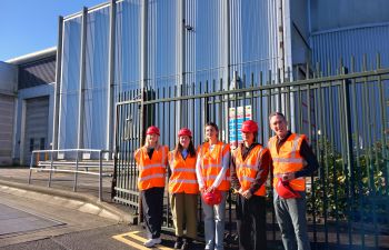 Sussex staff and Community Ambassadors stand in front of the Materials Recovery Facility with high visibility jackets and helmets on