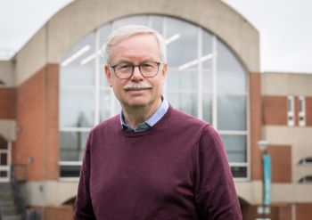 Dr Andrew Morgan wearing glasses and smiling, photographed standing in front of a red brick university of Sussex building