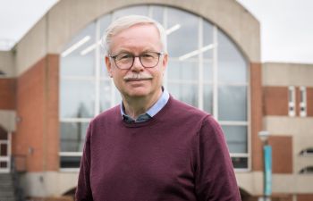 Dr Andrew Morgan wearing glasses and smiling, photographed standing in front of a red brick university of Sussex building