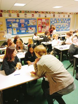 Young students seated at desks in a classroom, with a female teacher leaning down talking to two students at the front of class