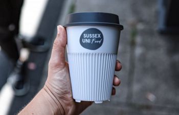 A caucasian woman's hand holding a returnable cup with a label that says 'Sussex Uni Food'