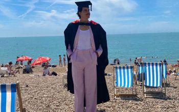 Pablo Santana in graduation clothing on the Brighton Beach with deck chairs and people in the background