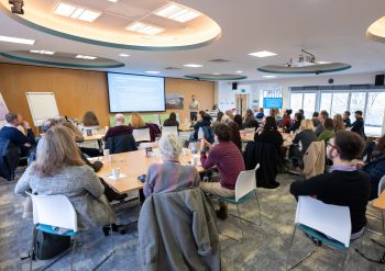 A group of people in a room with windows looking at a powerpoint presentation on a screen (pic from behind)