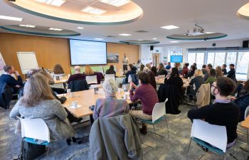 A group of people in a room with windows looking at a powerpoint presentation on a screen (pic from behind)