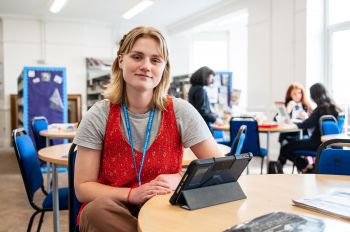 A young, female teacher trainee is seated smiling directly into the camera in a school setting. She has a laptop on the desk in front of her.