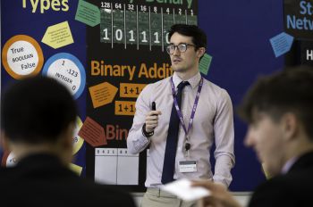 A young, male trainee teacher stands in front of maths symbols in a classroom setting