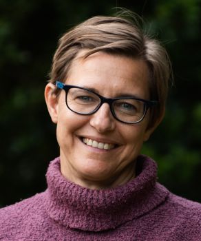 Head shot of woman with short blonde hair wearing glasses and a purple jumper and smiling to camera.