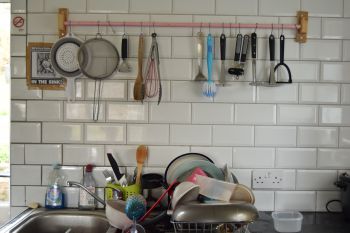 Kitchen sink sirrounded by white tiles with plates and a wine glass draining and kitchen utensils hanging above the sink on a pink rail.