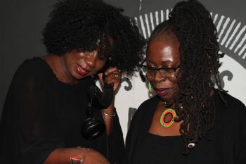 A photograph of two women looking down at some DJ decks (out of shot) one woman holds a wired headphone to her ear.