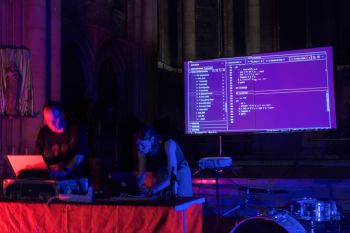 A photograph of a dark room bathed in blue and red light. Two people are stood over laptops on a table with a screen projected with code on a black background behind them. In the bottom right of the photo a large drum set sits underneath the screen..