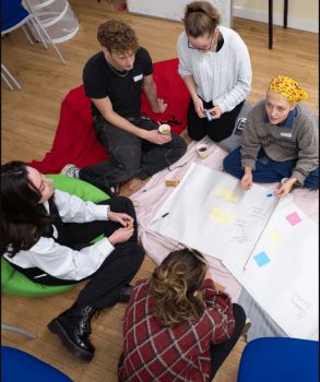 5 students sat around in a circle on the floor, discussing a project & mapping onto a large white sheet of paper with some colourful post-its on it.