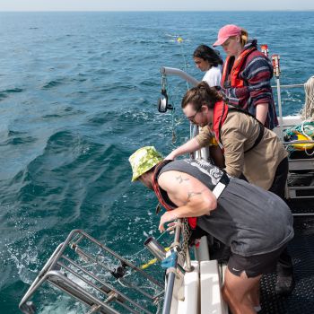 Four people leaning over the side of a boat with scientific equipment to scoop up seawater and measure it for eDNA