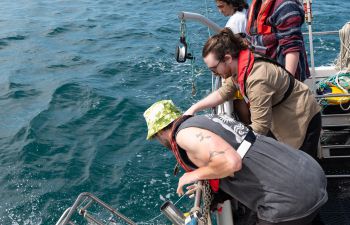 Four people leaning over the side of a boat with scientific equipment to scoop up seawater and measure it for eDNA