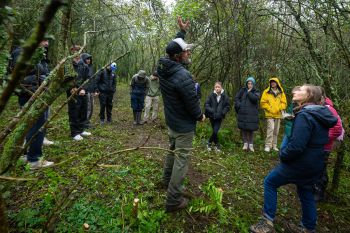 A group of students in a shrubby clearing surrounding by trees, looking at the teacher (a man in a cap) pointing and explaining