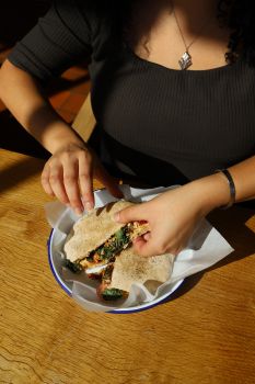 You can see the torso of a woman wearing a black top who's tearing into a healthy-looking flatbread; indoors but with the sun shining on her