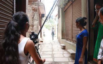 Photograph of the back of a teenage girl standing behind a camera on a tripod, to her right, three girls stand on a doorstep looking into the distance. She is shooting an image of the residential alleyway that they are stood in.