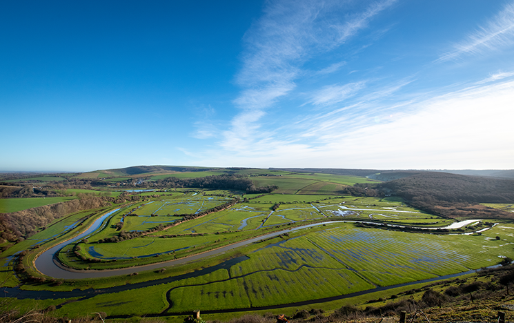 An aerial shot of Cuckmere Haven in Sussex: vast, open green land with a river winding through in the foreground and blue sky above