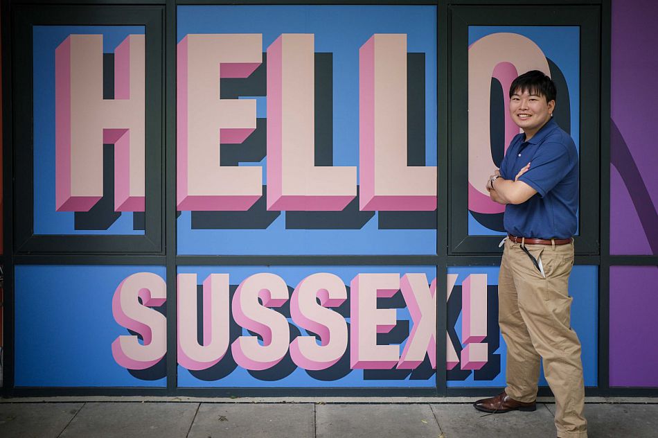 Takuto stood in front of a sign that reads 'Hello Sussex'