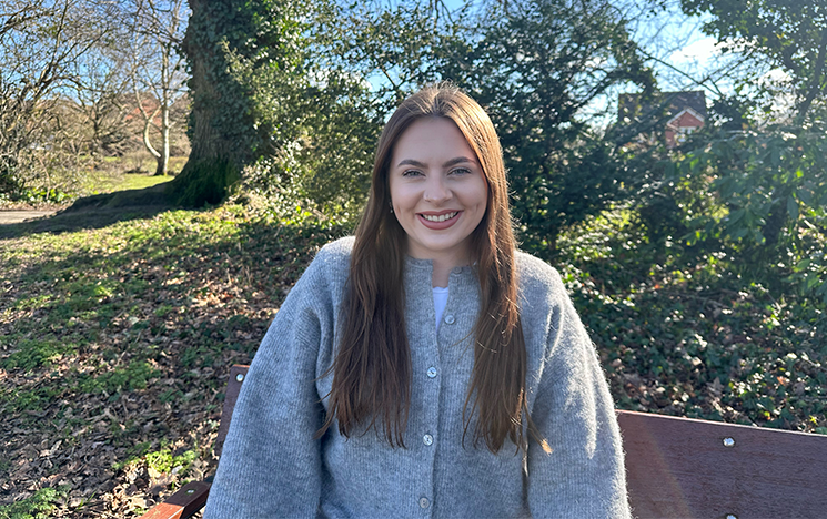 A student in a grey cardigan standing in a green space with trees in the background.