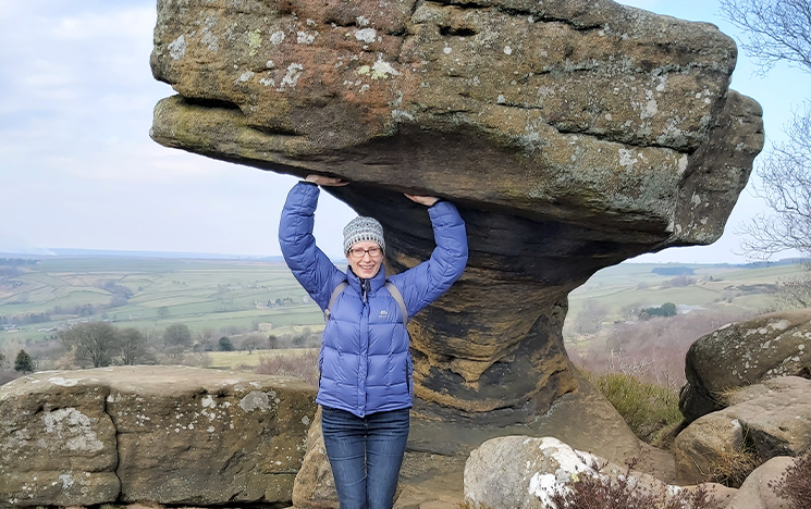 A woman in a coat and jeans, standing underneath a rock, raising her arms to make it look like she's holding it up.