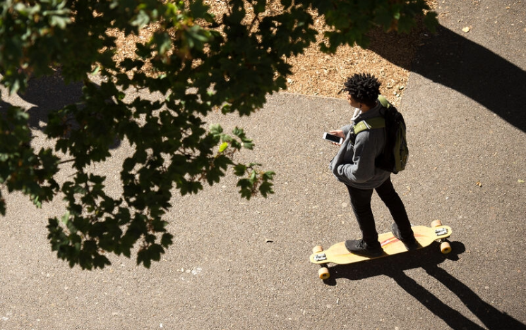 A student riding a skateboard on a sunlit paved path on campus while looking at a smartphone, with tree branches casting shadows.