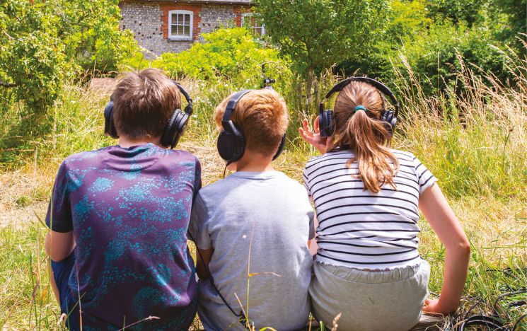 The backs of three children with earphones sitting on some grass in a row