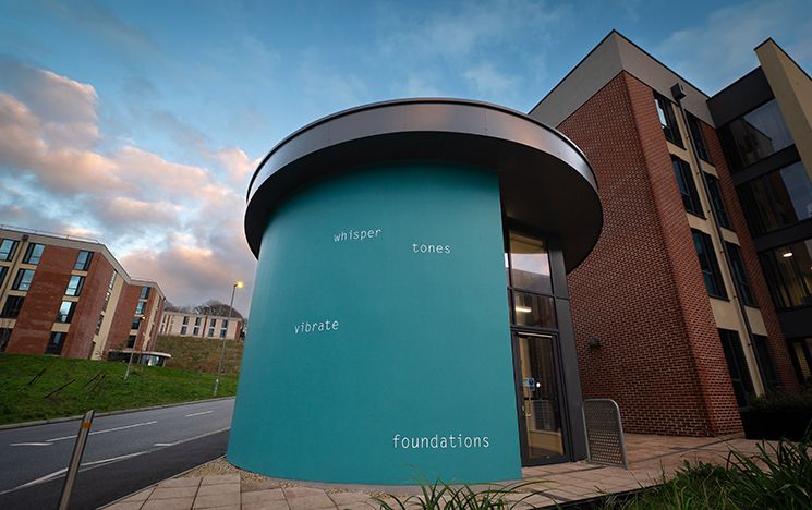 Helen Cammock's public artwork on campus, a curved teal wall with the words 'whisper tones vibrate foundations' in white text spaced randomly
