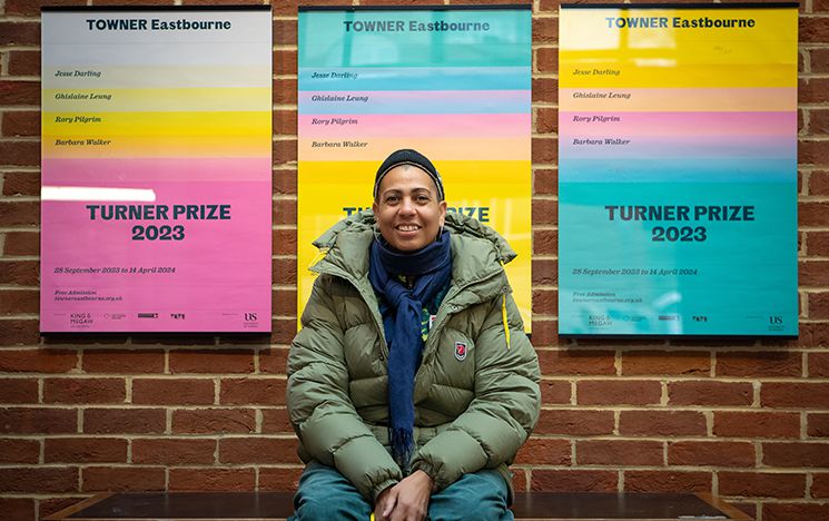 Helen Cammock sitting on a bench seat in ACCA with Turner Prize at Towner Eastbourne posters behind her