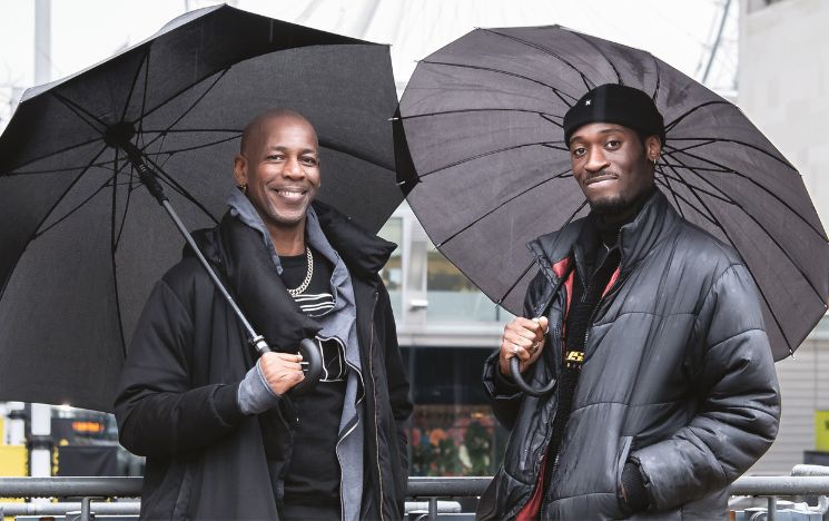 Topher Campbell and Jordi M. Carter stood together holding umbrellas on a rainy day in London.