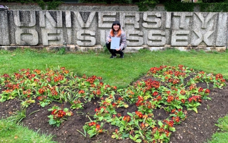 Huyen Le holding a copy of their dissertation in front of one of the University of Sussex concrete signs on campus.