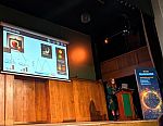 Dr Jenny Boston presenting at a New Scientist event in September 2024. Behind Jenny on stage is a slide on colour matching with images illustrating her points, including a red apple in front of a TV showing said apple.