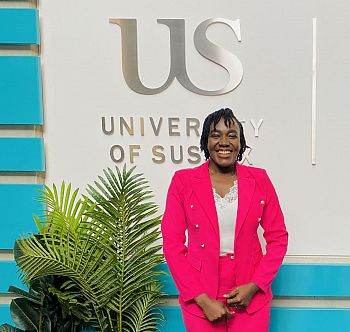 Charity Akanam standing in front of the University of Sussex logo. There is green fern plants next to her. Photo credit Charity Akanam,