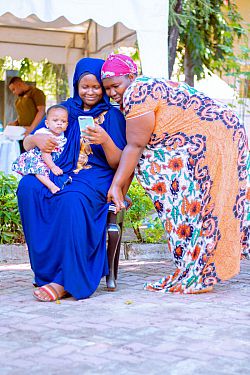  Two women looking at a phone. One is sat holding a baby while the other leans on the chair.