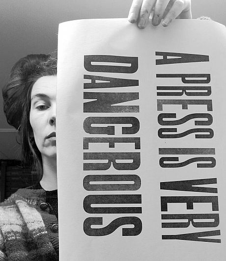 Person holds a sign saying 'the press is very dangerous'. The sign covers half their face. They are looking straight into the camera with a serious expression