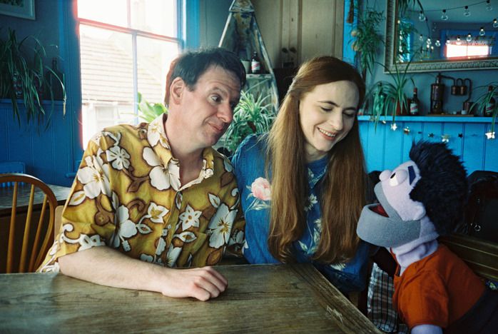 A man and a woman sit at a table looking happily at a male puppet.