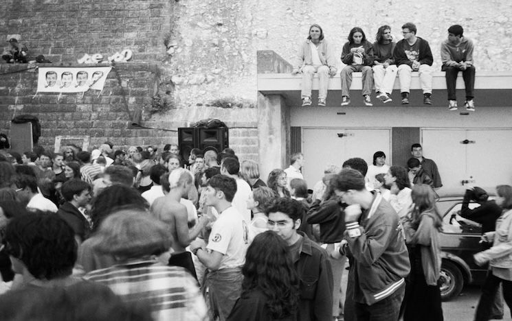 Black and white photograph of a crowd of young people gathered by a sound system on Undercliff Walk. Some people sit on top of the roof of old beachuts.