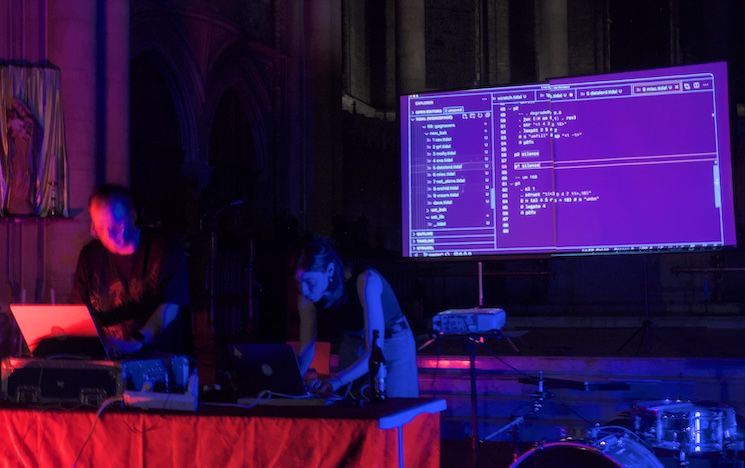 Photo of two people on laptops with a screen behind them showing lines of code next to a drum kit.