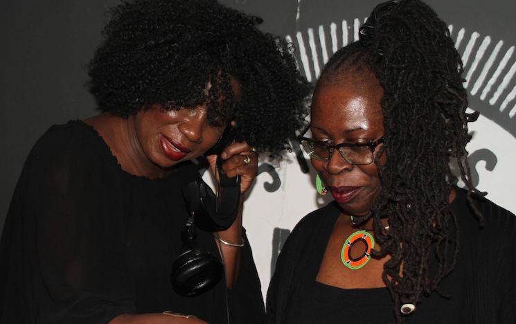 Image of two women (Nzinga Sounds) looking down at DJ decks. One woman is holding some headphones to her ear.