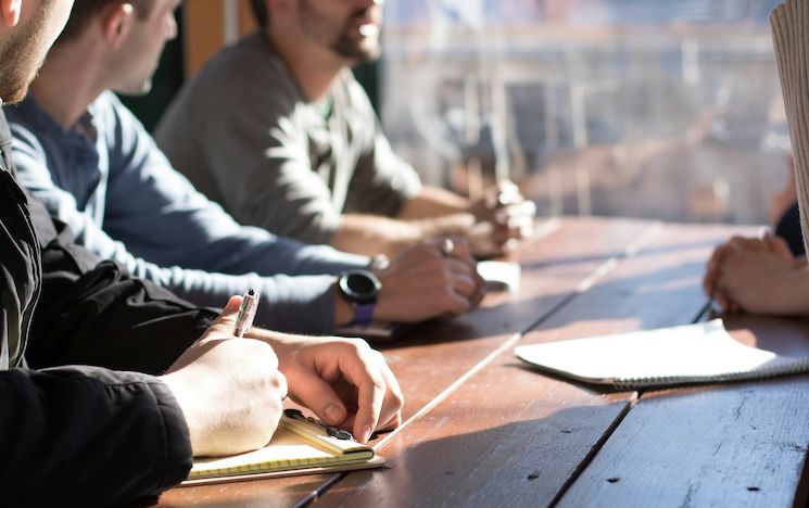 photo of people sitting on chair in front of table while holding pens during daytime