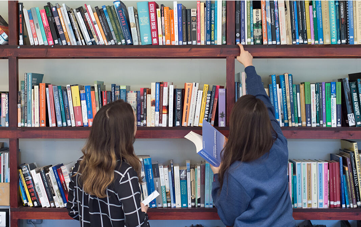 Two students with their backs to the camera choosing books in a library.