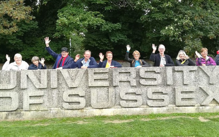A group of alumni standing behind and leaning on the top of the concrete University of Sussex sign on campus