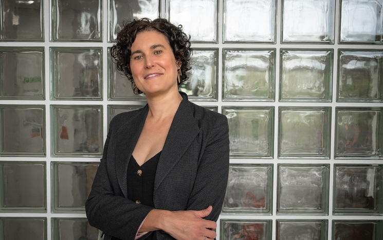 Dr Maya Semrau smiling with her arms folded in front of a glass brick wall