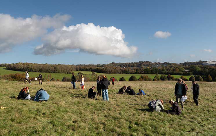 A group of students conducting fieldwork in the grassy South Downs. Some are sitting or
kneeling on the grass while others stand in small groups, engaged in observation and
discussion.