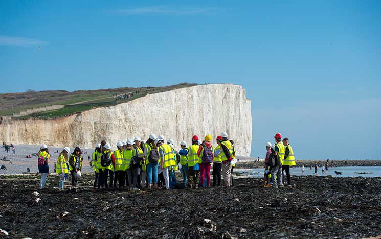 A group of students conducting fieldwork wearing high-visibility vests and hard hats.
They are gathered on a rocky shoreline near the base of the Seven Sisters cliffs.