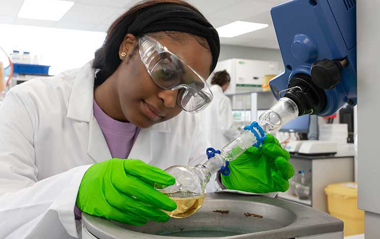 A student in a white lab coat, green gloves, and safety goggles conducting a laboratory
experiment.