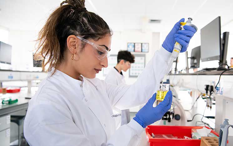 A student in a white lab coat, blue gloves, and safety goggles looking conducting a
laboratory experiment.