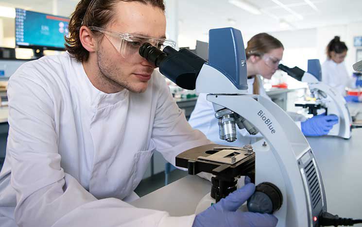 A student in a white lab coat, blue gloves, and safety goggles looking through a
microscope in a laboratory.