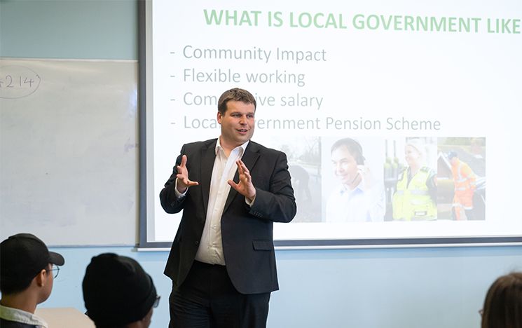 A man in a suit delivering a presentation to students, with a slide projection in the background saying 'What is local government like?'