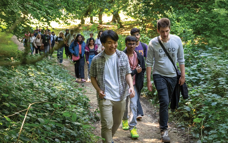 A large group of smiling people walking together along a footpath through green leafy woods, one person wearing a top with the words Good Life on